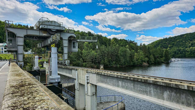 An industrial view of a hydraulic structure against a backdrop of wooded hills and a river. The dam's structural elements and lifting mechanisms are visible in the foreground. - Powered by Adobe