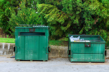 Two green dumpsters sit side by side outside, surrounded by trees grass on sunny day.