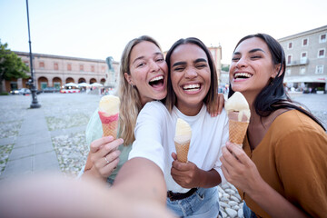 Three cheerful female friends are enjoying a summer day, taking a selfie while laughing and eating ice cream cones in a picturesque town square