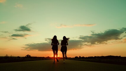 Athletic young women running along an asphalt road at sunset, leading healthy fitness lifestyle. Girls are training, running along road, teamwork of athletes. Slow motion. People run sun together, sky
