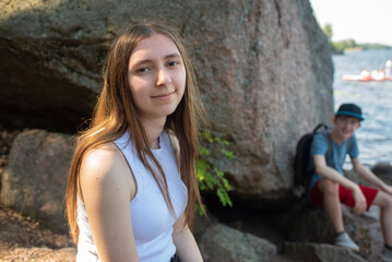 A teenage girl or young woman in a white T-shirt and jeans sits on a rock on the river bank.