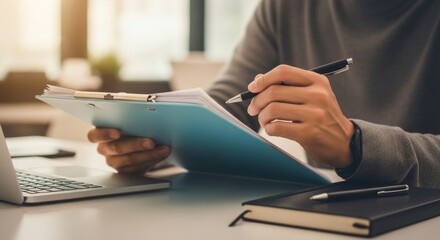 A person reviews documents on a clipboard while working at a desk with a laptop and notebook