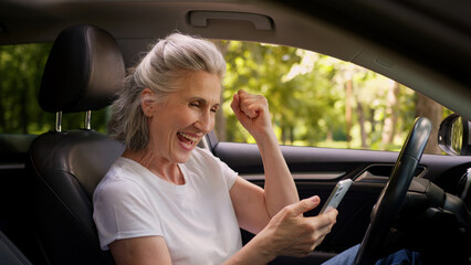 Naklejka premium Cheerful Caucasian mature woman elderly senior female retired pensioner car automobile driver inside auto using mobile phone playing game clench fist celebrating win victory betting success offer deal