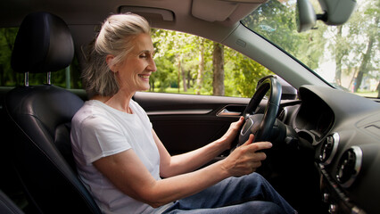 Excited overjoyed Caucasian smiling woman lady senior elderly aged female rental service client car happy driver in modern electric automobile admiring salon stroke steering wheel buying new transport