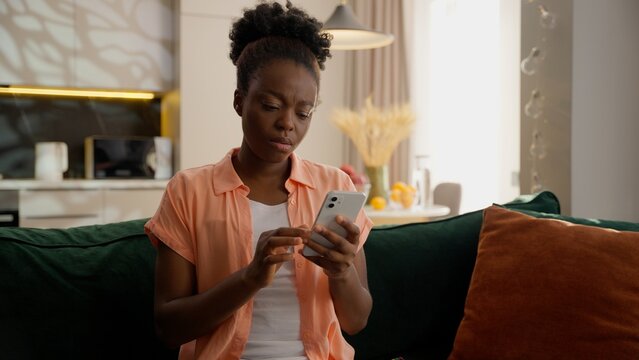 Confused unhappy sad ethnic African American business woman female girl businesswoman depressed client homeowner user holding mobile phone using app betting gambling fail lost loss money scam at home
