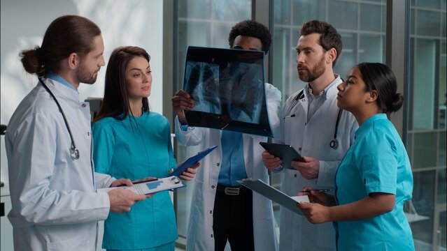 Group of five multiethnic medical students interns men women doctors analyzing patient x-ray result using digital tablet writing clipboard consulting discuss health treatment inside hospital hallway