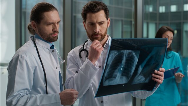 Two Caucasian males men doctors interns talking about patient result x-ray image lungs surgeon and therapist radiologist consulting discussion diagnosis fluoroscopy speaking indoors hospital hallway