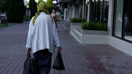 Elderly woman walking on city sidewalk carrying bags in warm evening light