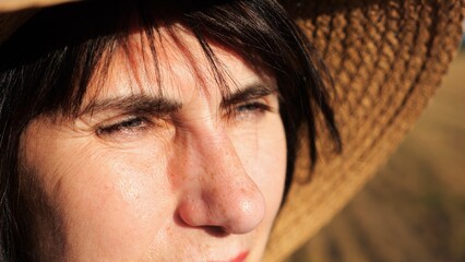 Female farmer in straw hat looking into wheat field and examining plantation at sunset. Close up face of adult beautiful woman illuminating by bright rays of sun. Concept of agriculture business