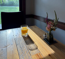 A glass of orange juice on a table with flowers and morning light coming from a window. 