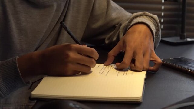Close-up of hands writing in a notebook with a pen. Focus on study, work, journaling, or creative process on a dark desk.