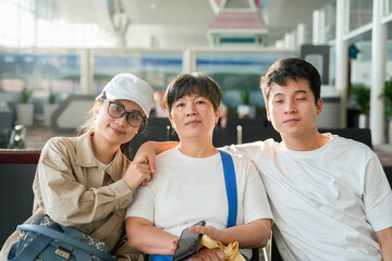 Group of three Asian people, likely a family, sitting together in an airport waiting area. Focus on travel, family, and group portrait