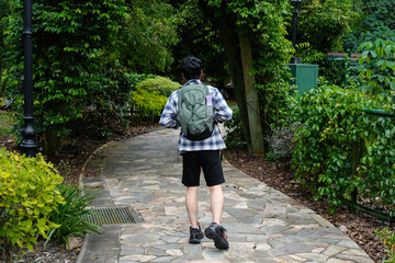 Fototapeta premium Young man with a backpack walking on a winding path in a lush, green park or garden. Focus on nature, outdoor activity, and solo exploration.
