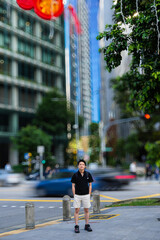 Fototapeta premium young man standing on a street corner in an urban area. He is wearing a black shirt, light-colored shorts, a backpack.Traffic lights, moving cars creating motion blur