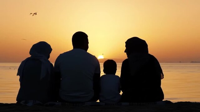 Family sitting on the sand watching sunset, captured as silhouettes. Wide static frame with colorful sky. Calm, emotional, and peaceful family vacation scene.
