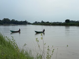 Two Asian men row separate wooden boats across the calm river on a cloudy day
