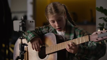 Caucasian artist little girl singer playing guitar recording mobile phone blog camera tripod blogger child influencer kid play music song watching online video lesson musical instrument skills at home
