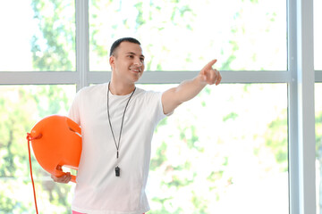 Happy male lifeguard with life board and whistle pointing at something near window in pool
