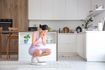 Shocked young Asian woman measuring weight on scales in kitchen. Diet concept