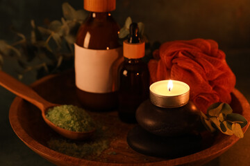 Wooden tray, spa stones, burning candle, spoon with sea salt and bottles of cosmetic product on black background, closeup
