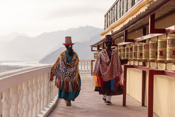 Two women in traditional Indian attire walk along a monastery in Leh, Ladakh, beside a row of prayer wheels, surrounded by stunning Himalayan mountains in the soft morning light.