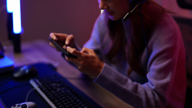Asian young adult woman streamer gamer playing mobile video game online using headset technology equipment in dark room glowing lights at night home setup