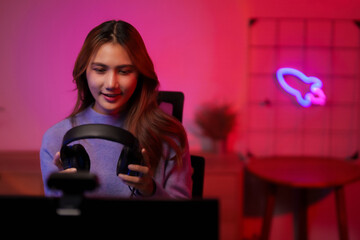 Young Asian adult woman streamer gamer preparing headphones for online video gaming and streaming technology setup at home room with neon light