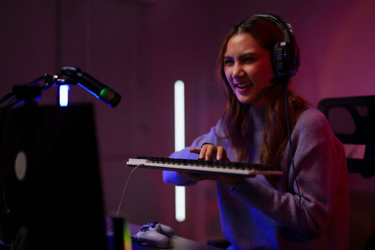 Young Asian woman online streamer gamer plays video game using headset keyboard computer setup dark room neon lighting esports competition gaming lifestyle content creator