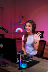 Young Asian adult female streamer live streaming from home studio gaming setup waving engaging audience online technology