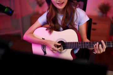 Passionate young adult Asian woman musician playing acoustic guitar streaming music online broadcasting entertaining content creative home studio pink light atmosphere
