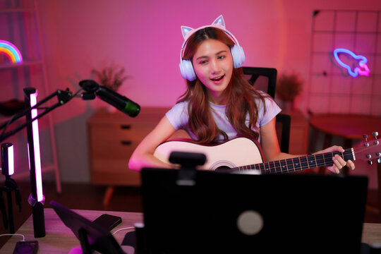 Asian young adult woman playing acoustic guitar live streaming music performance online creating content from vibrant home studio with technology equipment