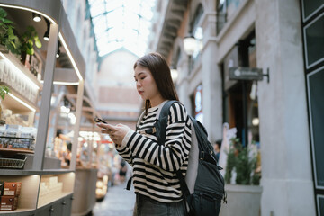 Fototapeta premium A modern young woman in a striped shirt stands in an urban mall, holding a laptop and smartphone, embodying a tech-savvy, stylish, and connected digital lifestyle. 