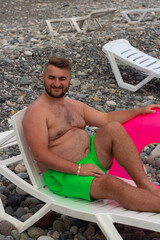 Vertical travel photo. Full body young adult tanned fat man with beard, big belly, green shorts, magenta swim ring sits on white plastic sun chaise lounge on pebble sea coast. Summer rest, trip