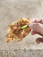 Close-up shot of the right hand holding fried fritters with green chilies. A crispy fried food made from a mixture of flour and carrots, bean sprouts, and cabbage. Indonesian food with negative space.