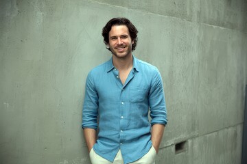 A smiling man in a blue linen shirt stands against a grey wall