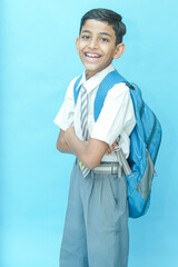 Happy Indian schoolboy wearing uniform and bag standing isolated over blue background, Education...