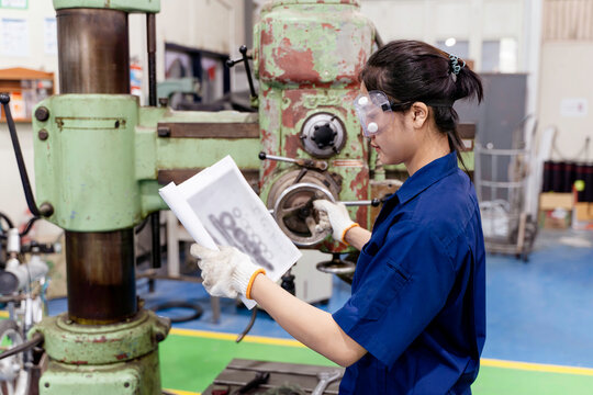 Asian woman Industrial engineer in uniform holds a sample print of different sized gears that will be manufactured into parts on a lathe and drill press at industrial workshop. - Powered by Adobe