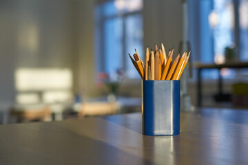 Pencils in blue holder on table in sunlit room near windows.