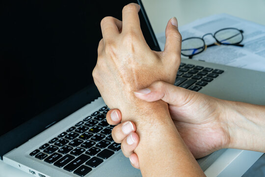 Asian man experiencing wrist pain from office syndrome, seated at a cluttered desk with laptop and papers&mdash;highlighting ergonomic strain in a modern workspace. Office syndrome concept.