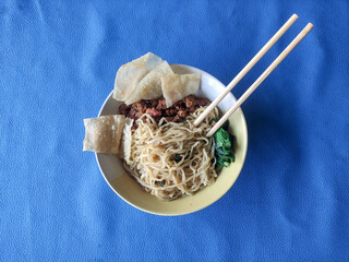 A bowl of chicken noodles with dumplings and green vegetables on a blue tablecloth