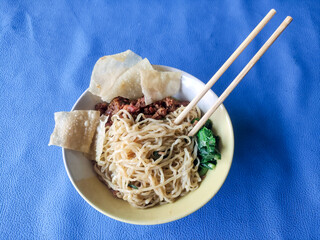 A bowl of chicken noodles with dumplings and green vegetables on a blue tablecloth