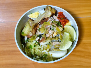 A bowl of Indonesian chicken soto with shredded chicken, sliced boiled egg, vermicelli, sliced cabbage, bean sprouts, celery, chili sauce and lime, mixed with delicious sauce. On a wooden table.