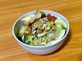 A bowl of Indonesian chicken soto with shredded chicken, sliced boiled egg, vermicelli, sliced cabbage, bean sprouts, celery, chili sauce and lime, mixed with delicious sauce. On a wooden table.