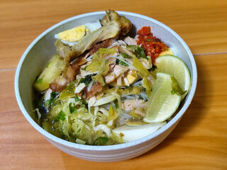 A bowl of Indonesian chicken soto with shredded chicken, sliced boiled egg, vermicelli, sliced cabbage, bean sprouts, celery, chili sauce and lime, mixed with delicious sauce. On a wooden table.