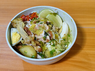 A bowl of Indonesian chicken soto with shredded chicken, sliced boiled egg, vermicelli, sliced cabbage, bean sprouts, celery, chili sauce and lime, mixed with delicious sauce. On a wooden table.