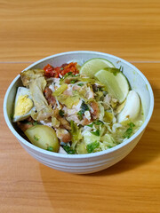 A bowl of Indonesian chicken soto with shredded chicken, sliced boiled egg, vermicelli, sliced cabbage, bean sprouts, celery, chili sauce and lime, mixed with delicious sauce. On a wooden table.