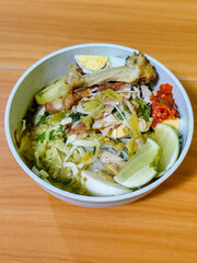 A bowl of Indonesian chicken soto with shredded chicken, sliced boiled egg, vermicelli, sliced cabbage, bean sprouts, celery, chili sauce and lime, mixed with delicious sauce. On a wooden table.
