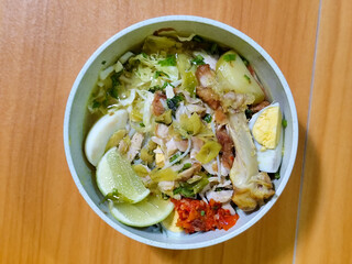 A bowl of Indonesian chicken soto with shredded chicken, sliced boiled egg, vermicelli, sliced cabbage, bean sprouts, celery, chili sauce and lime, mixed with delicious sauce. On a wooden table.