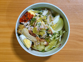 A bowl of Indonesian chicken soto with shredded chicken, sliced boiled egg, vermicelli, sliced cabbage, bean sprouts, celery, chili sauce and lime, mixed with delicious sauce. On a wooden table.