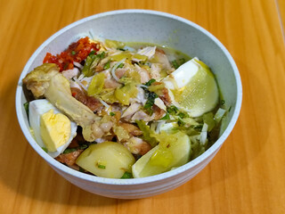 A bowl of Indonesian chicken soto with shredded chicken, sliced boiled egg, vermicelli, sliced cabbage, bean sprouts, celery, chili sauce and lime, mixed with delicious sauce. On a wooden table.